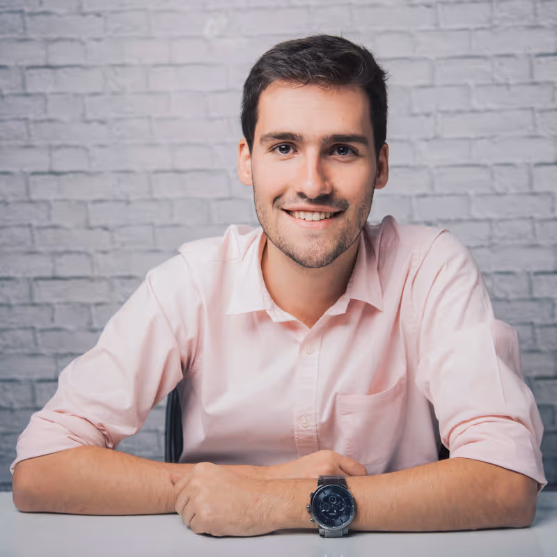 Young man with dark hair wearing a light pink shirt and a wristwatch, smiling while sitting at a table with a gray brick wall background.