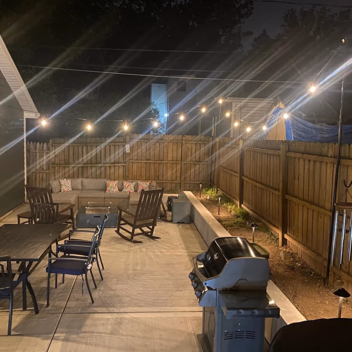 Nighttime backyard patio with string lights overhead, outdoor dining table with chairs, rocking chairs, a sectional sofa with colorful pillows, and a grill.