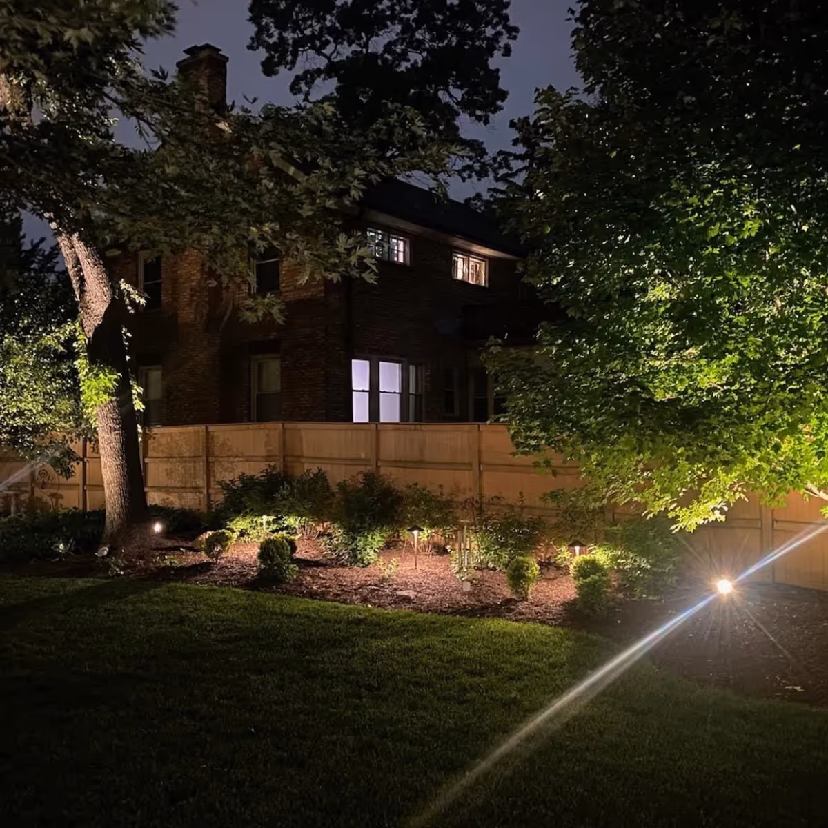 Nighttime view of a garden with outdoor landscape lighting illuminating trees, bushes, and a wooden fence in front of a brick house.