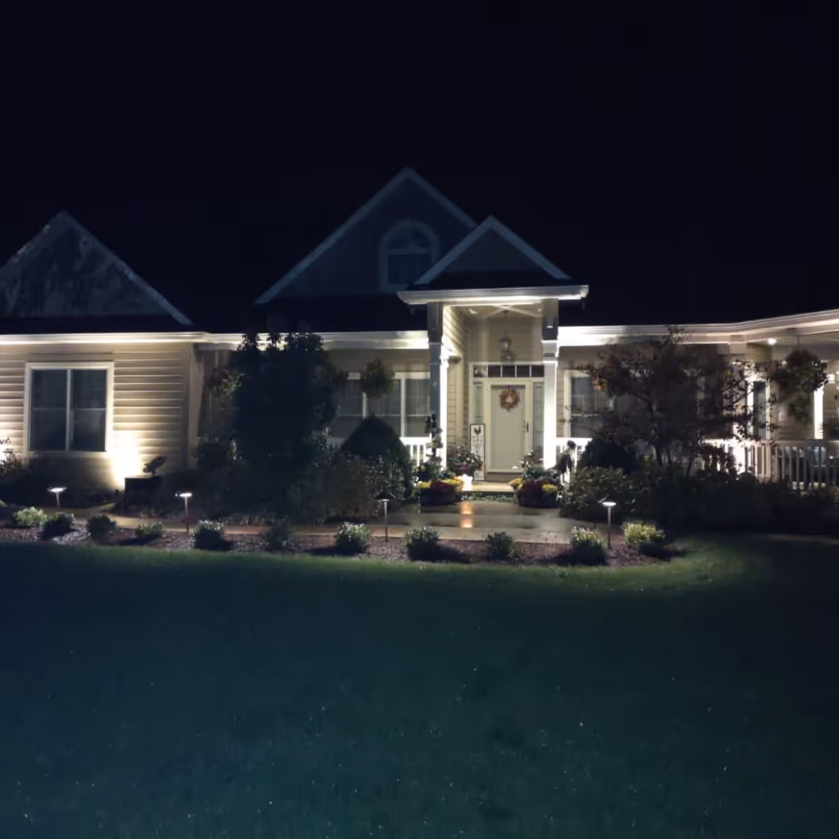 Front of a house illuminated by outdoor landscape and porch lighting at night, with bushes and plants lining the walkway.