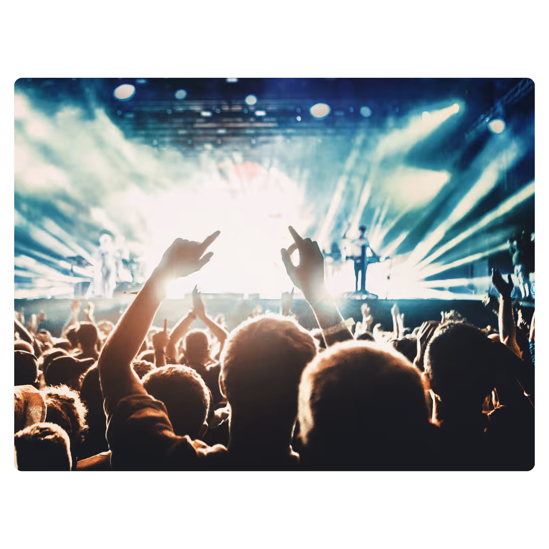 Concert goers dancing with raised hands in front of a stage with bright lights.