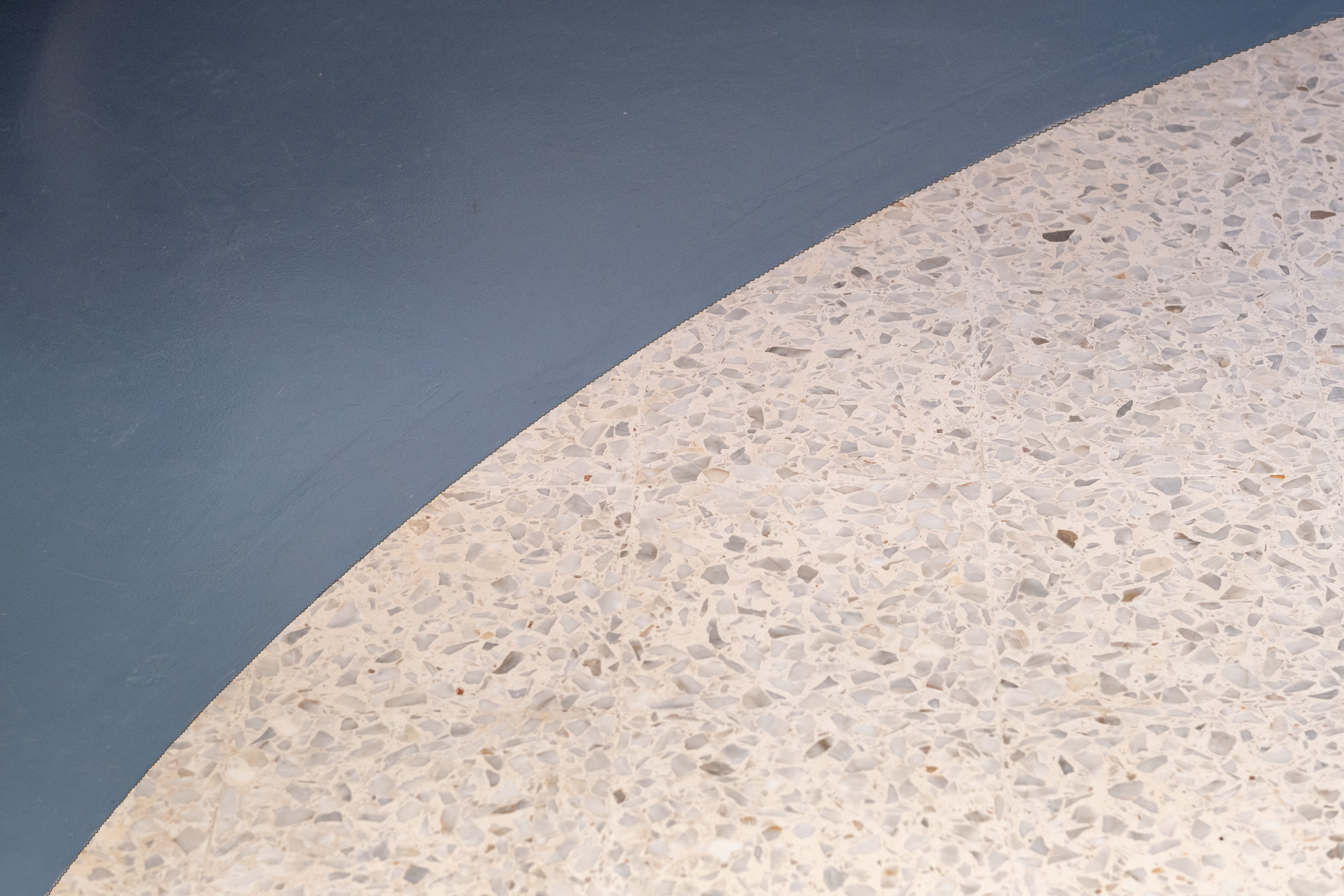 La nuova sede operativa dello studio nasce da un progetto di ristrutturazione volto a preservare l’identità originaria dell’immobile, valorizzandone le qualità architettoniche e adattandole a un ambiente di lavoro contemporaneo.
Materiali storici e proporzioni originarie vengono rispettati: il pavimento in graniglia restaurato racconta la storia dello spazio, mentre grandi finestre e soffitti alti garantiscono luce e apertura.
Le esigenze tecniche diventano spunti progettuali: gli impianti sono integrati in un inserto in resina blu che segue l’arco architettonico e conferisce continuità allo spazio. La stessa finitura si ritrova nella meeting room e nell’ufficio privato, accanto all’open space pensato per stimolare collaborazione e concentrazione.
Il mobilio su misura introduce materiali caldi e superfici tattili, in contrasto armonioso con i toni neutri. Una libreria in legno separa la zona operativa dalla waiting area e dalla materioteca, cuore della ricerca progettuale.
Il risultato è uno spazio essenziale e accogliente, in cui memoria e contemporaneità si fondono in modo coerente con la visione dello studio.