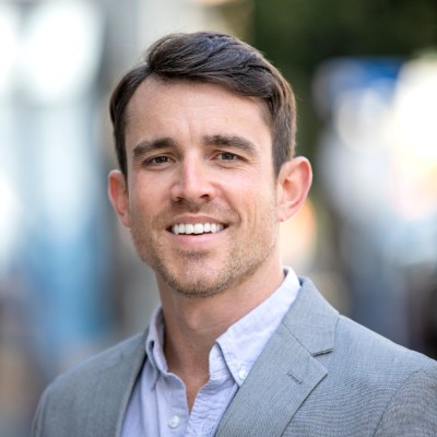 Smiling Matt Fellows wearing a light blue shirt and gray blazer, with a blurred outdoor background.