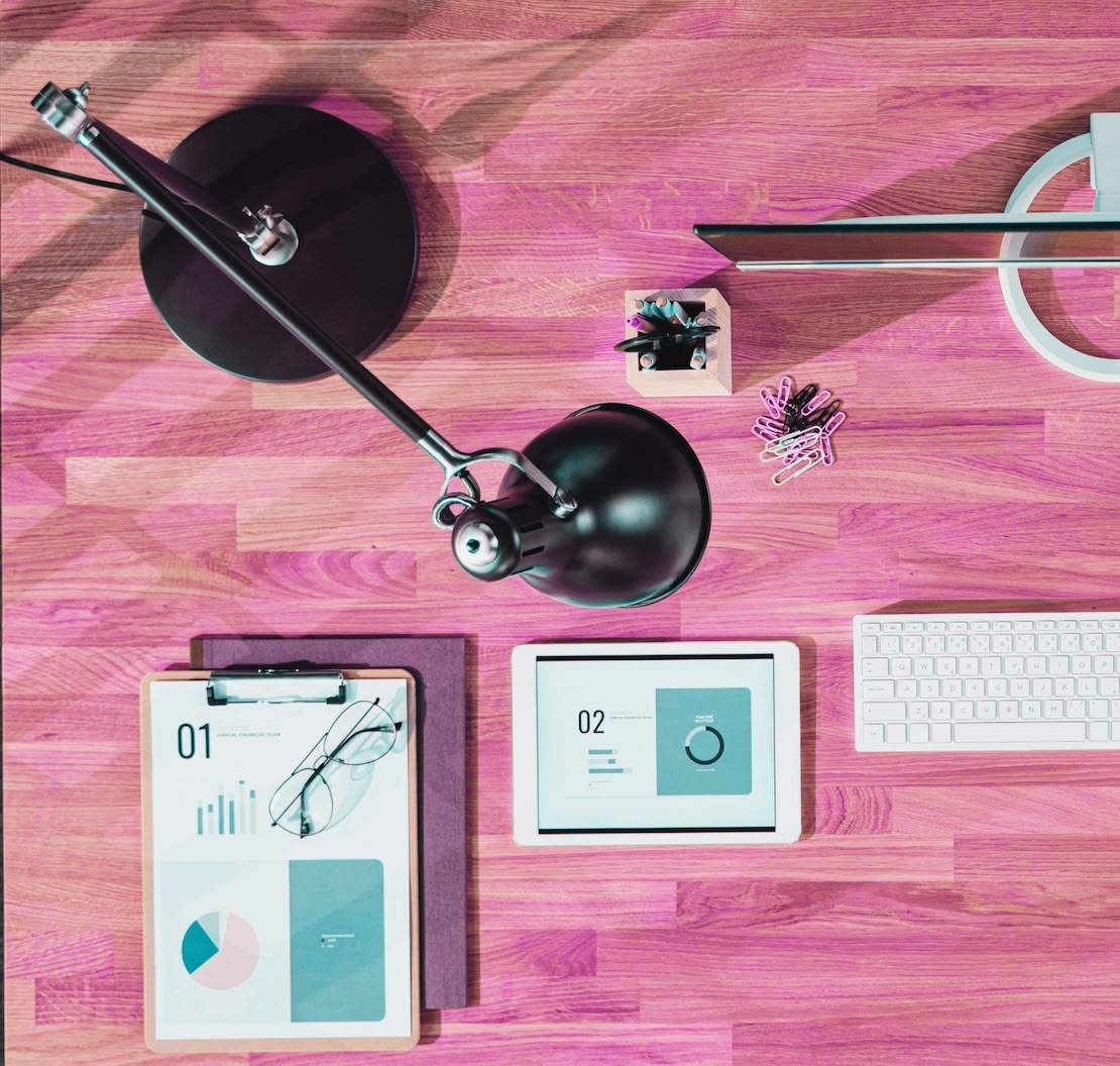 A wooden desk with a lamp, a clipboard, a tablet, a keyboard, paper clips, and a pen holder.