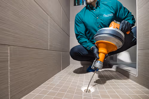 Person using a plumber's drain snake tool to unclog a shower drain inside a tiled shower.