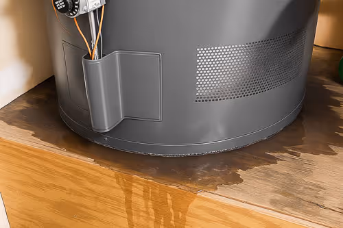 Base of a water heater with visible water leakage pooling on the wooden floor beneath it.