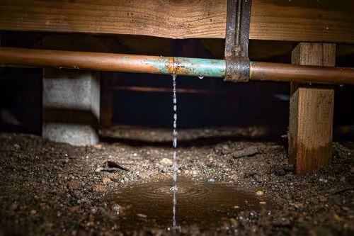 Water dripping from a corroded copper pipe under a wooden floor, forming a small puddle on dirt ground.