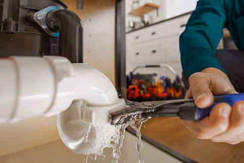 Person using a wrench to fix a leaking white kitchen pipe under a sink with water flowing out.