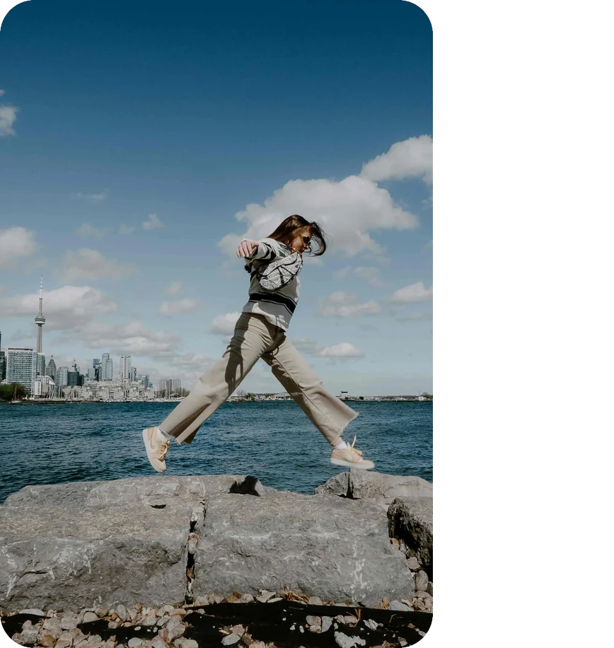 Woman leaping across waterfront rocks with the Toronto skyline and CN Tower behind her
