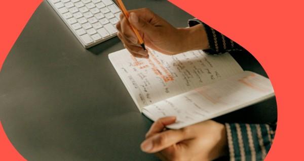 A person writing in a book with a computer keyboard in the background.