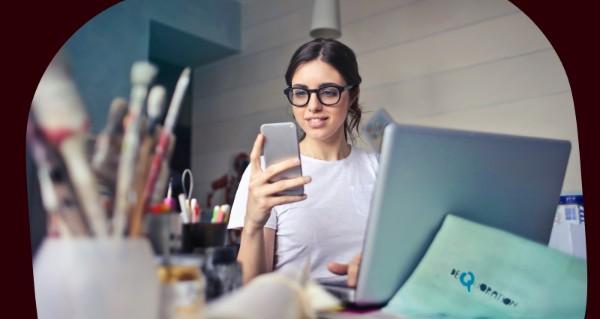 A woman holding a cell phone in front of a laptop.