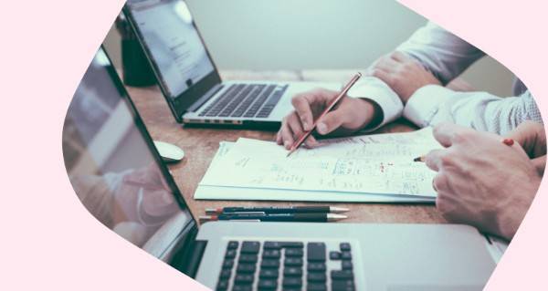 A man writing on a piece of paper with two laptops on a table.