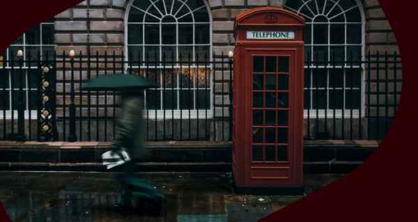A person walking with an umbrella in front of a red phone booth.