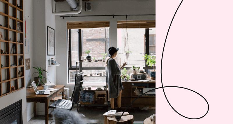A woman in a grey sweater stands in front of a window with a potted plant.