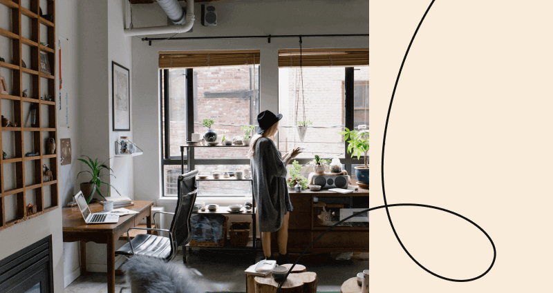 A woman in a grey sweater stands in a room with a window and plants.