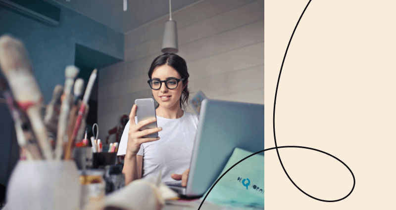 A woman is using a laptop and holding a cell phone.