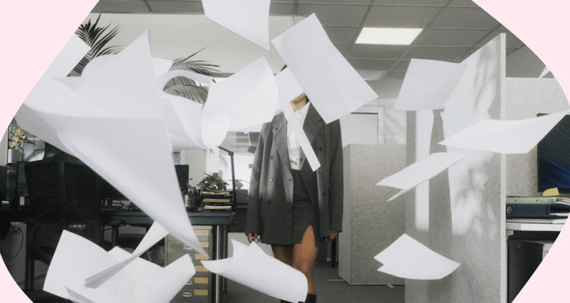 A woman in a grey coat stands in front of a desk with a bunch of paper flying around her.