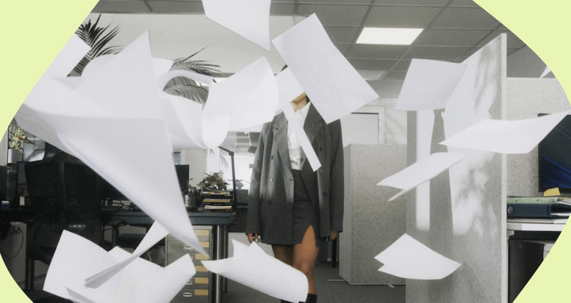 A woman in a grey suit stands in front of a desk with a bunch of paper flying around her.