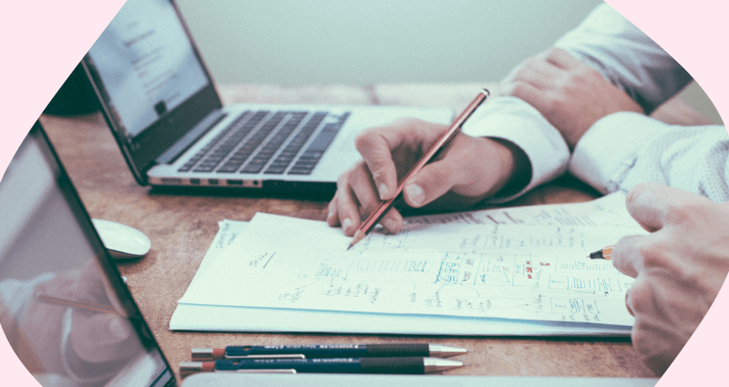 A person writing on a piece of paper with a laptop and pens in the background.