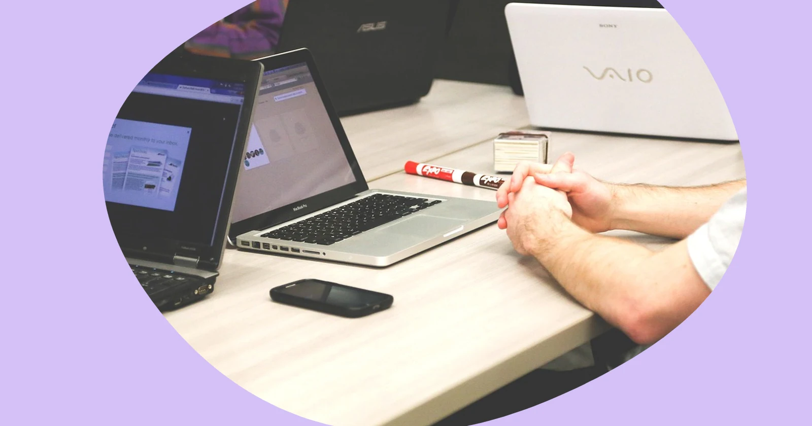 Hands clasped on a desk beside several open laptops, a smartphone and a marker.