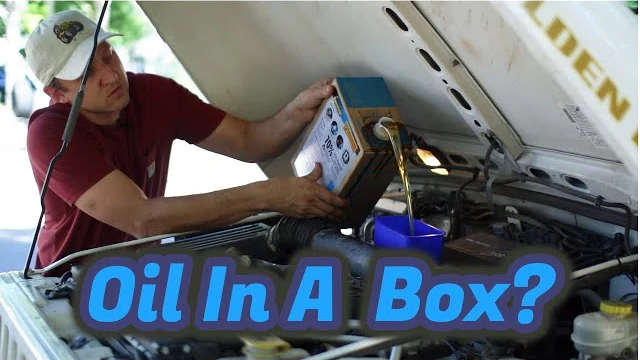 Man in a maroon shirt and cap pouring motor oil from a cardboard box into a car engine under the raised hood, with the text Oil In A Box? across the image