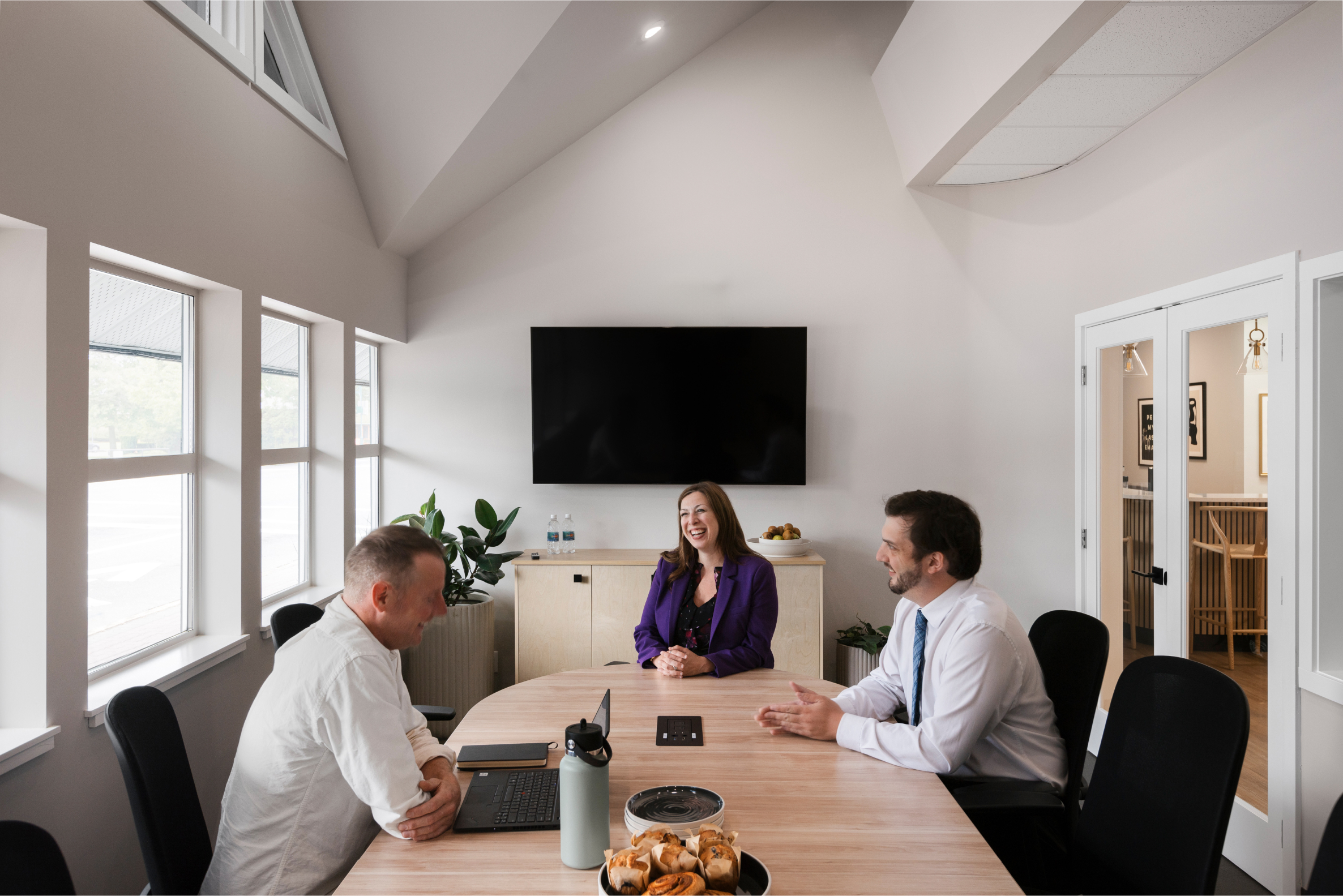 Three people sitting and smiling in conversation at a long board room table. In front of a conference call television