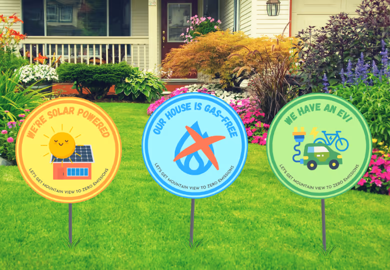 Three yard signs on green lawn in front of a house with garden flowers reading: 'We're solar powered,' 'Our house is gas-free,' and 'We have an EVI,' promoting zero emissions.