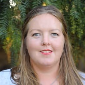 Portrait of a smiling woman with long light brown hair and blue eyes, standing outdoors with green foliage in the background.