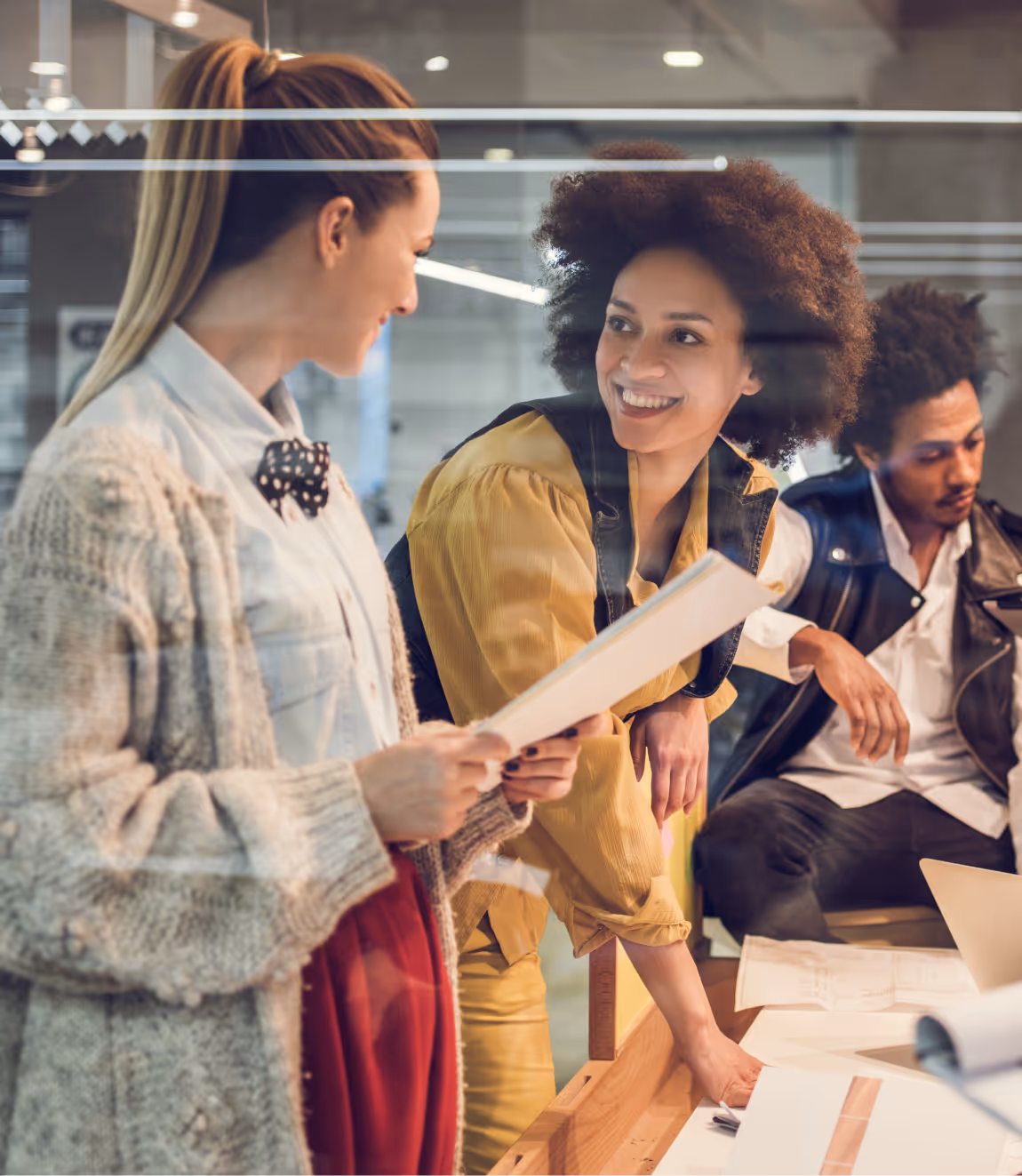 Three young professionals collaborating in an office, two women smiling and holding documents, one man focused on a laptop.