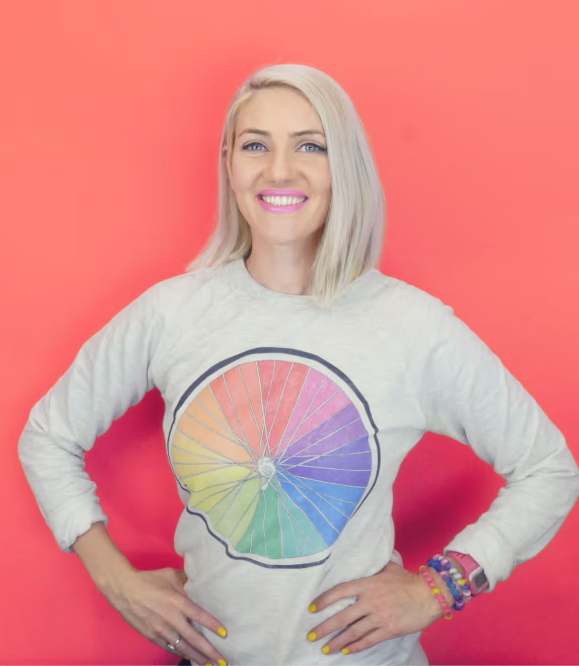 Smiling woman with blonde hair wearing a white sweatshirt with a colorful rainbow bicycle wheel design, standing against a pink background.