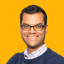 Smiling man with glasses wearing a navy sweater and checkered shirt on an orange background.