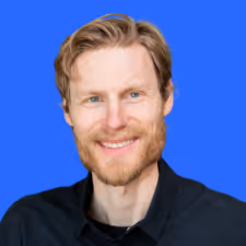 Smiling man with light brown hair and beard wearing a black shirt against a blue background.