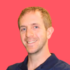 Smiling man with short light brown hair wearing a dark blue polo shirt against a solid pink background.