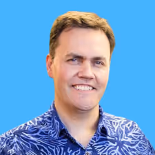 Smiling man with short brown hair wearing a blue patterned shirt against a bright blue background.