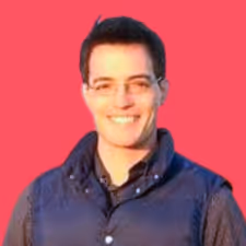 Smiling man with short dark hair and glasses wearing a dark vest and shirt against a pink background.