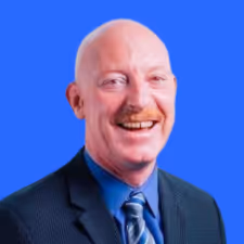 Smiling bald man with a mustache wearing a dark suit, blue shirt, and striped tie against a blue background.