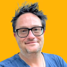 Smiling man with messy hair and black glasses wearing a blue shirt on an orange background.