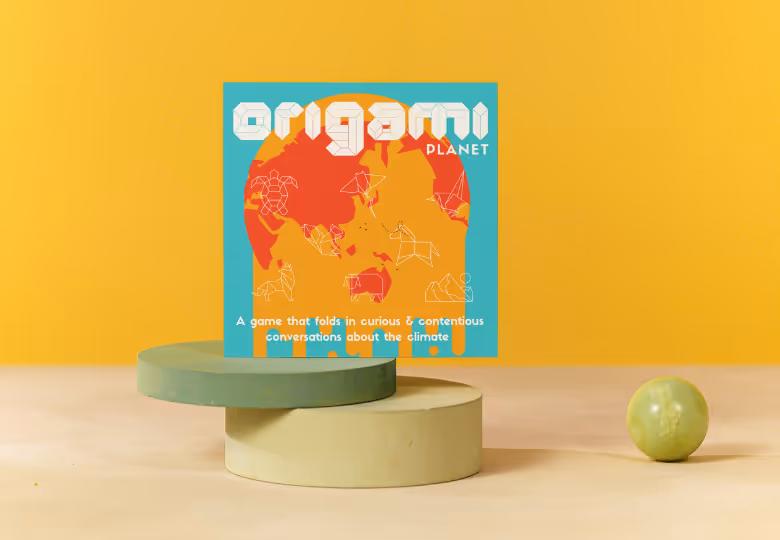 Origami Planet game box with a stylized orange map and white origami animal outlines on a turquoise background, placed on two stacked cylindrical platforms with a green marble on the side.