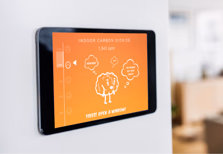 Digital display showing indoor carbon dioxide level at 1,042 ppm with an orange background, a brain cartoon thinking 'headache,' 'throb,' and 'I'm freakin' out over here!!!' and a message saying 'Yikes! Open a window!'