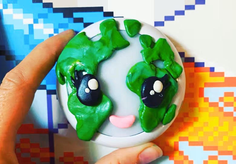 Hand holding a round object designed like Earth with large cartoon eyes and a pink smiling mouth.