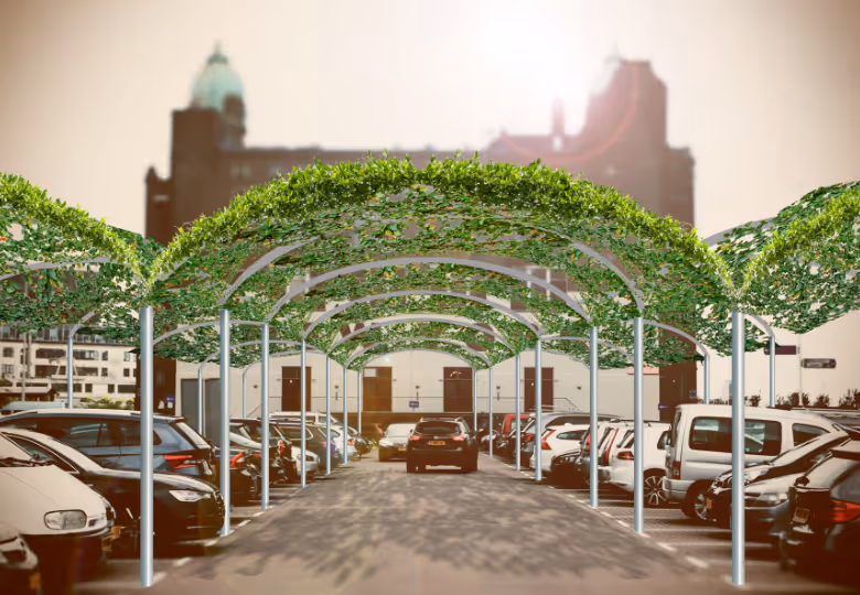 Outdoor parking lot with cars parked under metal arches covered in green leafy vines, with a city building in the background and sunlight visible.