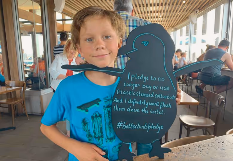 Young boy in a blue shirt holding a penguin-shaped sign with a pledge to avoid plastic stemmed cotton buds and not flush them, inside a busy cafe.