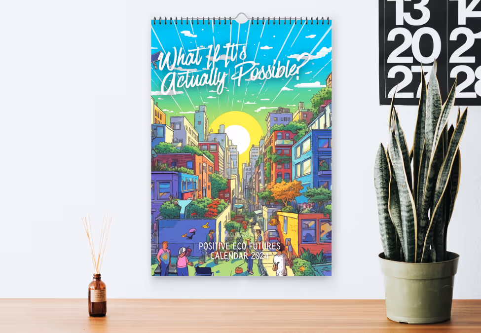 Wall calendar titled 'What If It's Actually Possible?' with colorful cityscape and greenery illustration, labeled 'Positive Eco Futures Calendar 2024', hanging above a wooden surface with a small brown diffuser and a green potted snake plant nearby.