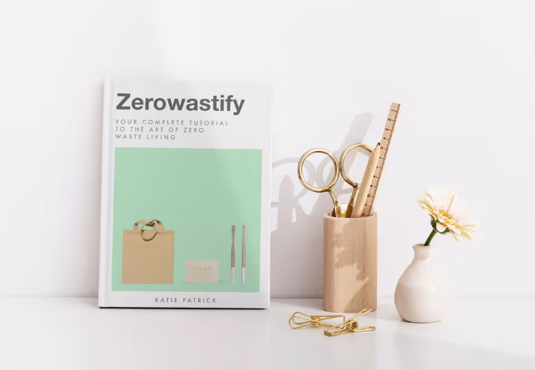 Book titled 'Zerowastify' by Katie Patrick beside a wooden holder with gold scissors, a ruler, a pen, and a small white vase with a single flower.