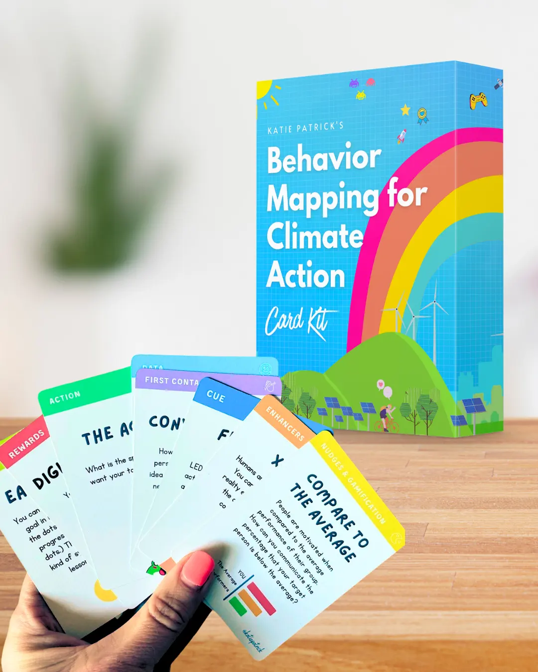 Hand holding a fan of colorful behavior mapping cards in front of a blue card kit box titled 'Behavior Mapping for Climate Action.'