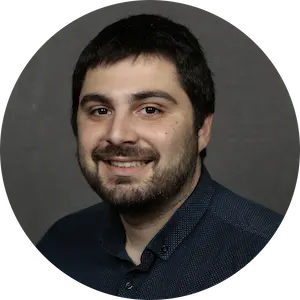 Smiling man with dark hair and beard wearing a dark blue shirt against a plain dark background.