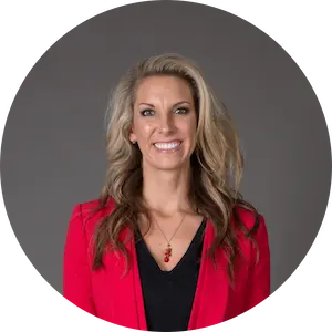 Smiling woman with long blonde hair wearing a red blazer and black top against a gray background.