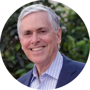 Smiling elderly man with gray hair wearing a blue blazer and striped shirt, standing outdoors with greenery in the background.