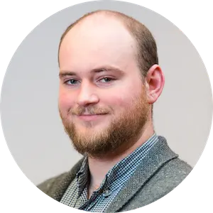 Smiling man with a beard and short hair wearing a patterned shirt and gray sweater against a neutral background.