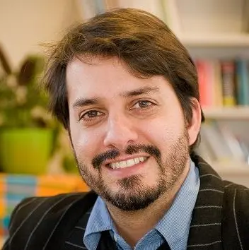 Smiling man with dark hair, beard, and mustache wearing a blue shirt and black pinstripe jacket indoors with blurred background.